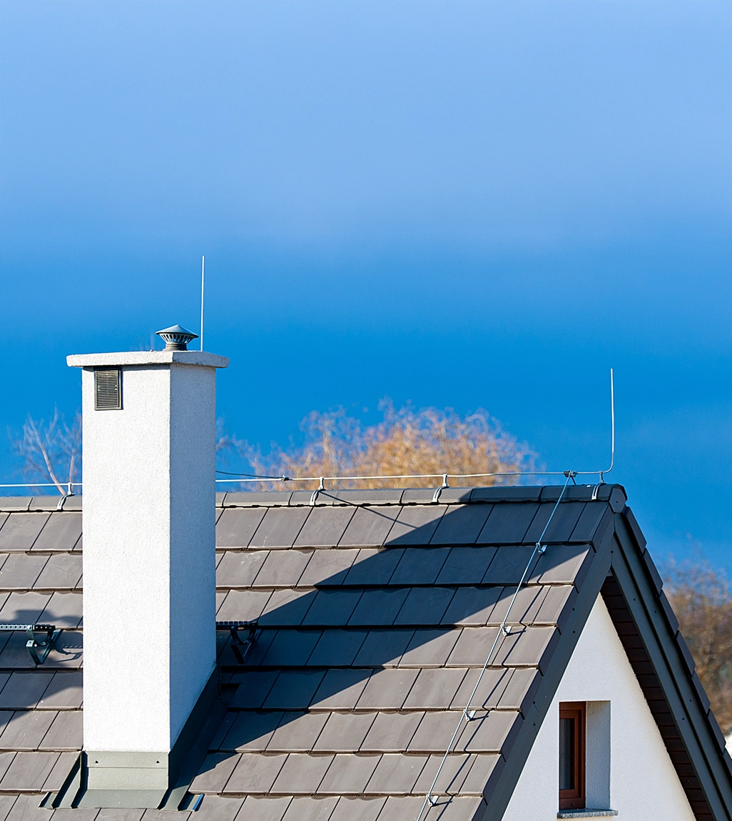 Modernes Wohnhaus mit installiertem Blitzschutzsystem auf dem Ziegeldach, sichtbar mit Blitzableitern, Erdungsleitungen und einem Schornstein bei klarem, blauem Himmel.