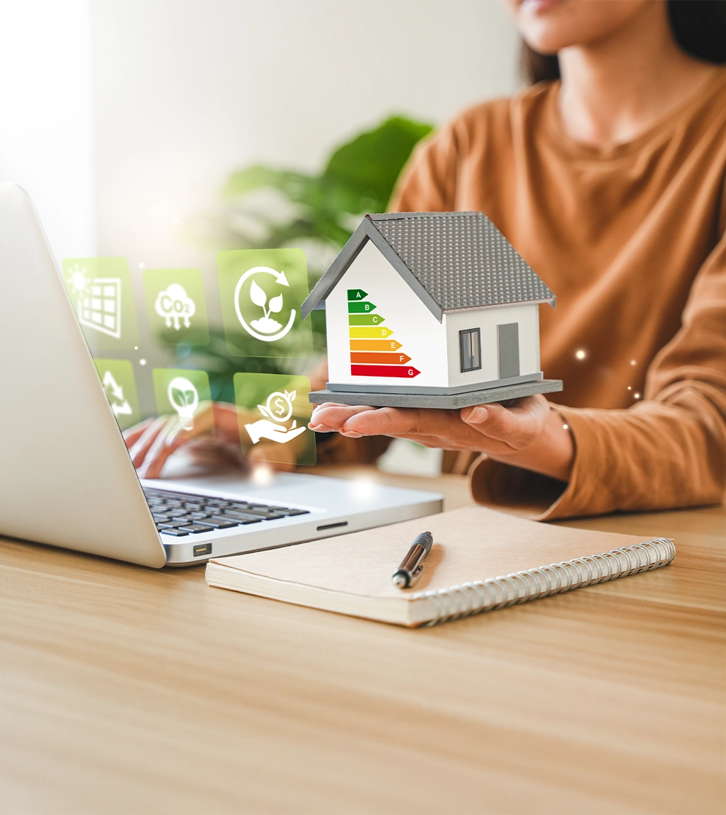 Frau hält ein digitales Modell eines Hauses mit Energieeffizienzklassen in der Hand, vor einem Laptop, umgeben von Icons zu Nachhaltigkeit, CO₂-Reduktion und Energieeinsparung.